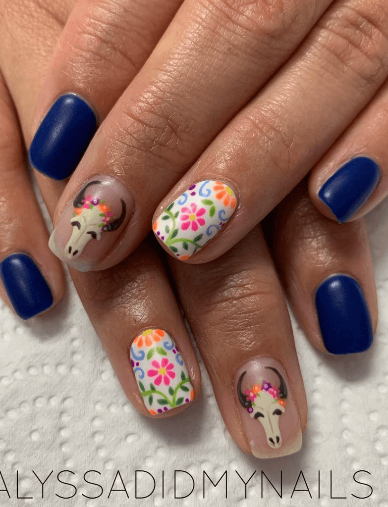 Nails with a mix of bold blue polish and clear bases featuring colorful floral designs and a bull skull, capturing the essence of Cinco de Mayo nails.