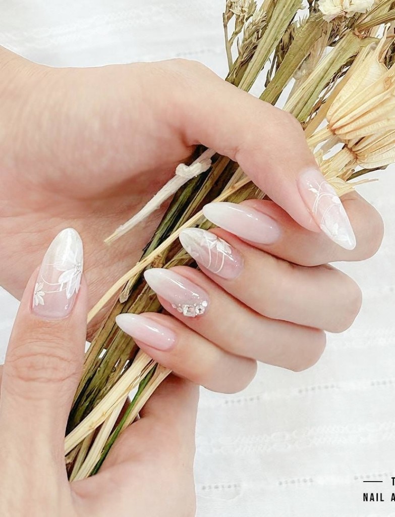 Almond-shaped wedding nails with a glossy translucent pink finish, featuring delicate white floral accents and a touch of rhinestone embellishments, harmonized with a natural dried bouquet, ready for a rustic chic wedding.