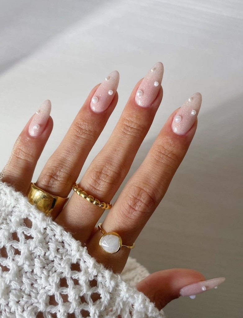 Long, almond-shaped wedding nails with a pearly pink base and subtle iridescent shimmer, each adorned with tiny white pearls, held elegantly against a crocheted white background.