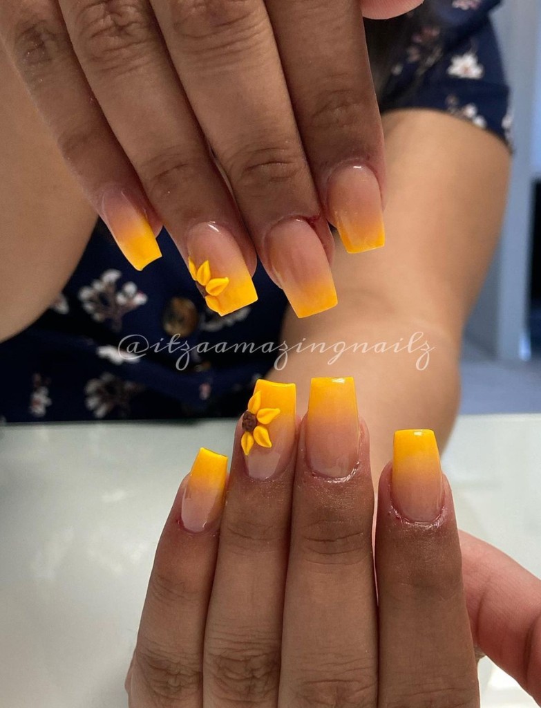 Hands with coffin-shaped nails featuring a yellow ombre effect, with delicate sunflower accents on select nails, shown above a blue floral patterned fabric.